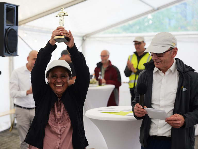 Eine Mitarbeiterin hält neben Geschäftsführer Ralf Zastrau eine Metallfigur in die Luft und blickt lachend in die Kamera bei der Jubiläumsfeier 20 Jahre Albertinen Services Hamburg ASH in Rellingen.