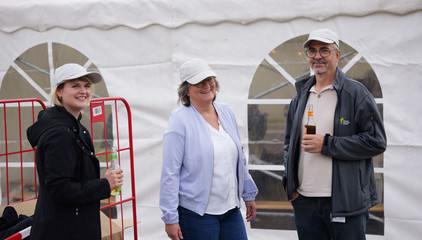 Drei Mitarbeitende mit Jubiläums-Kappen blicken vor dem Festzelt in die Kamera bei der Jubiläumsfeier 20 Jahre Albertinen Services Hamburg ASH in Rellingen.