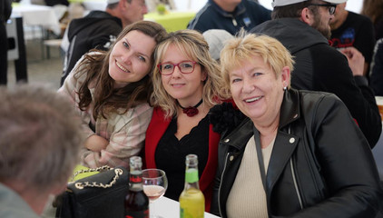 Drei Mitarbeiterinnen sitzen im Festzelt an einem Tisch und blicken lächelnd in die Kamera bei der Jubiläumsfeier 20 Jahre Albertinen Services Hamburg ASH in Rellingen.