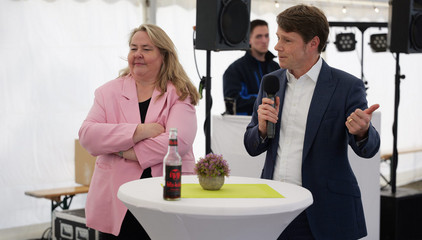 Konzerngeschäftsführer Dr. Thorsten Minuth spricht an einem Tisch neben Andrea Wichmann mit einem Mikrofon zu den Anwesenden im Festzelt bei der Jubiläumsfeier 20 Jahre Albertinen Services Hamburg ASH in Rellingen.